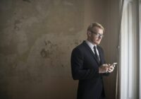 Side view of young distracted male entrepreneur in classy suit standing near window in shabby house and reading messages about eviction